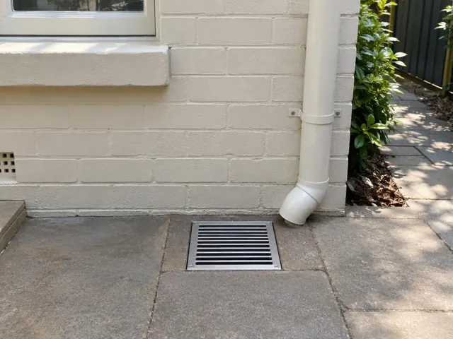 Outdoor gully grate set in paved area beside a house wall and downpipe in Chatswood