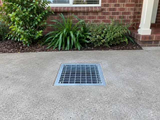 Stormwater grate seated in dry concrete driveway beside brick house and garden in Lane Cove