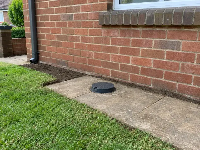 Capped sewer opening beside a brick house wall and paved path in Artarmon